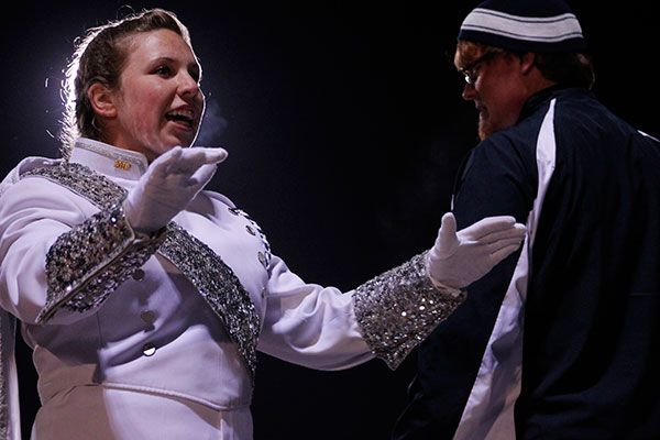 Uniformed marching band performers gesture to each other under stage lighting.