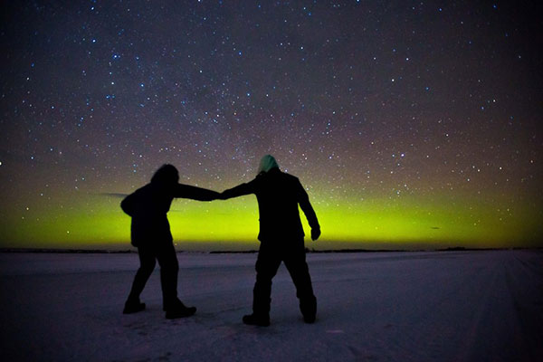 Two silhouetted figures hold hands beneath stars and green northern lights.