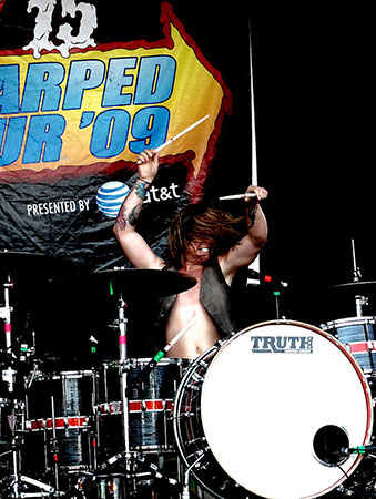 A shirtless drummer raises both sticks behind a drum kit during a live performance.