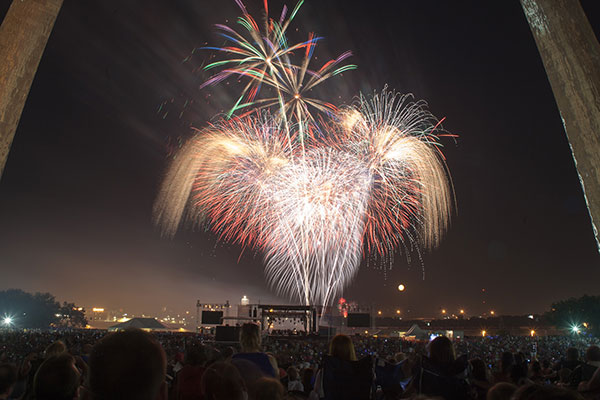 Colorful fireworks burst above a large nighttime crowd gathered near a distant stage.