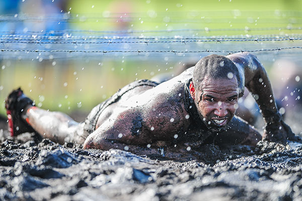 Mud-covered competitor crawls beneath barbed wire while water droplets fill the air.
