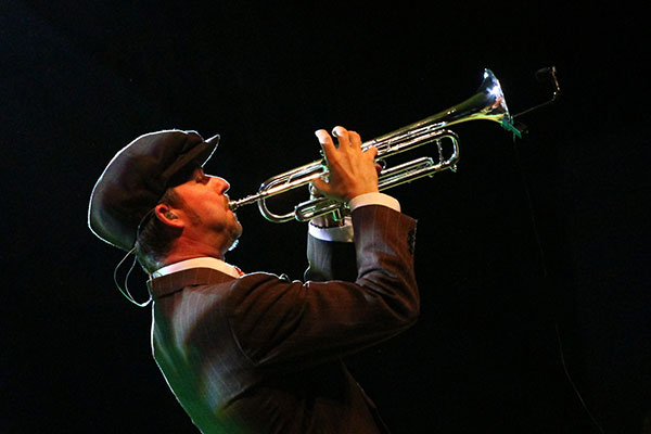Trumpet player in a dark suit and cap performs under stage lighting.
