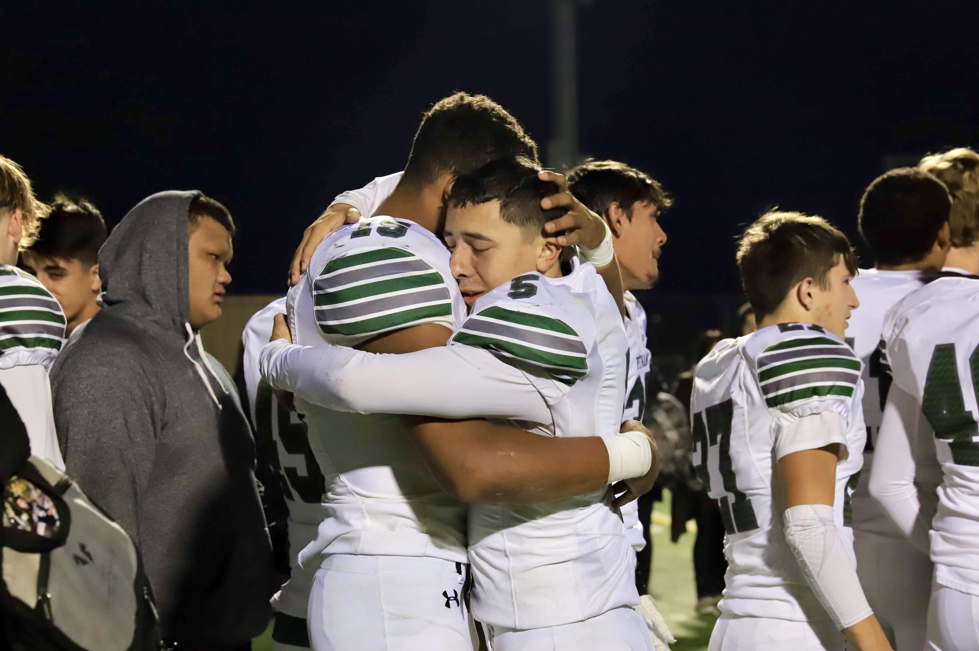 Two football players embrace on the field as teammates gather around them after a game.