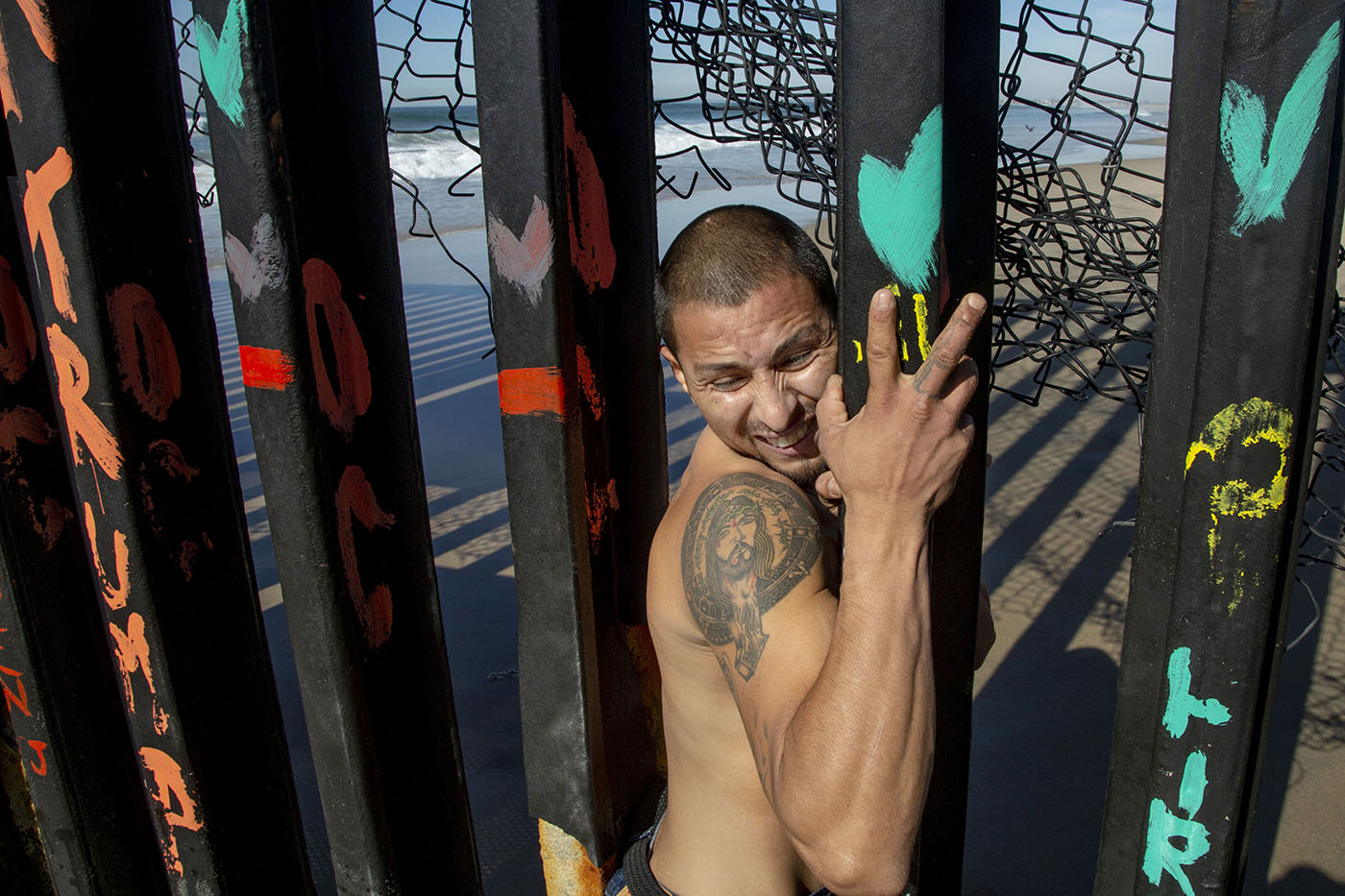 Shirtless tattooed man squeezes between painted metal border bars topped with tangled wire.