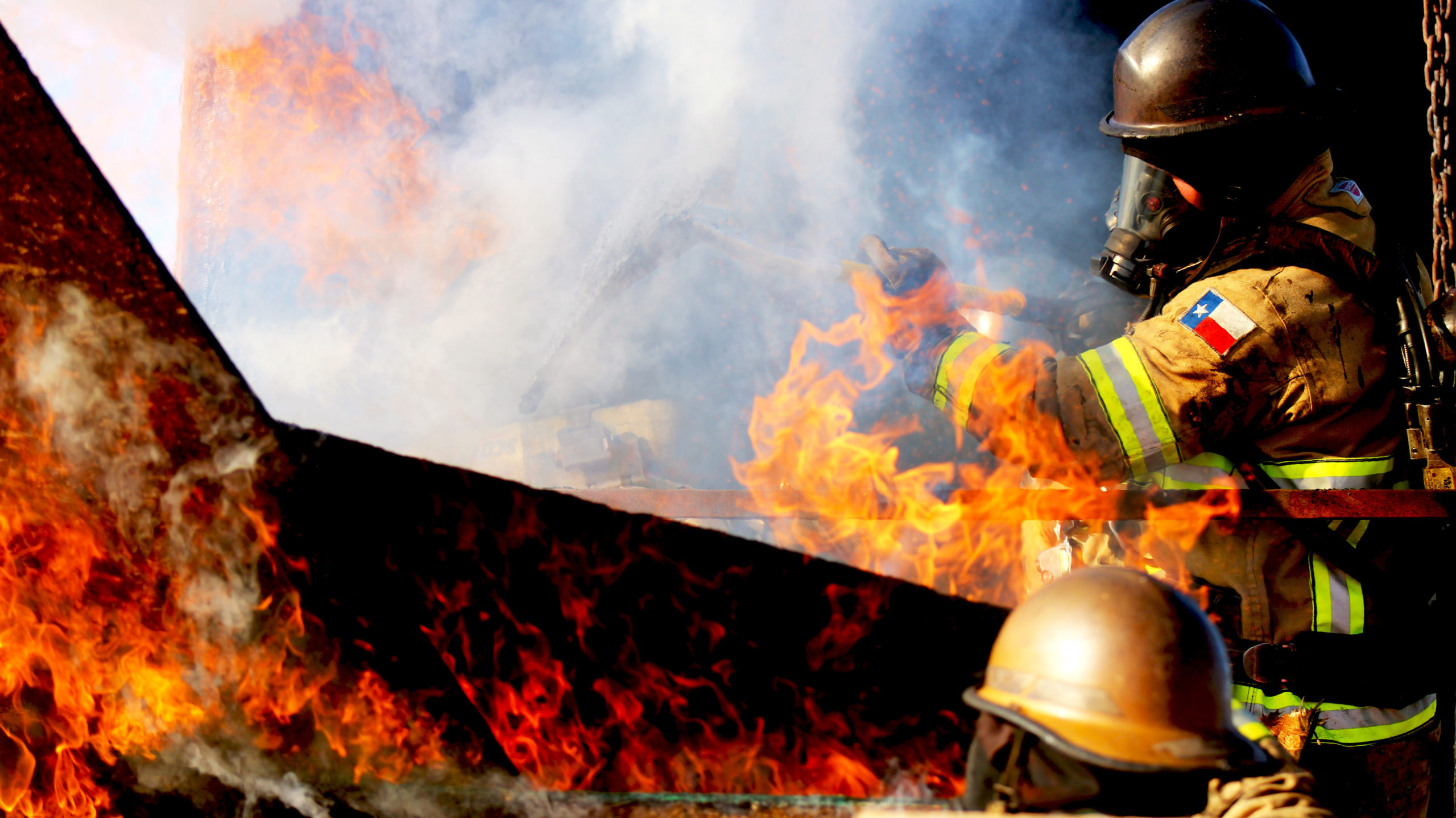Firefighter in protective gear works amid bright flames and thick smoke.