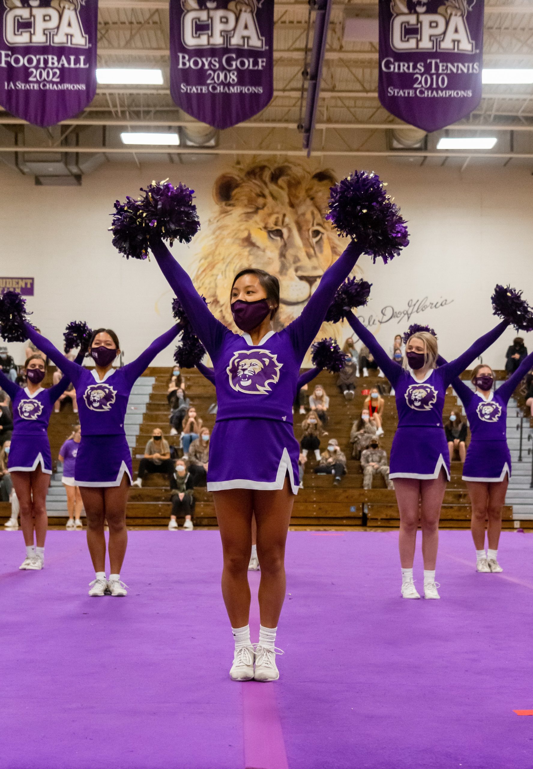 Masked cheerleaders in purple uniforms hold pom-poms overhead on a gymnasium floor.