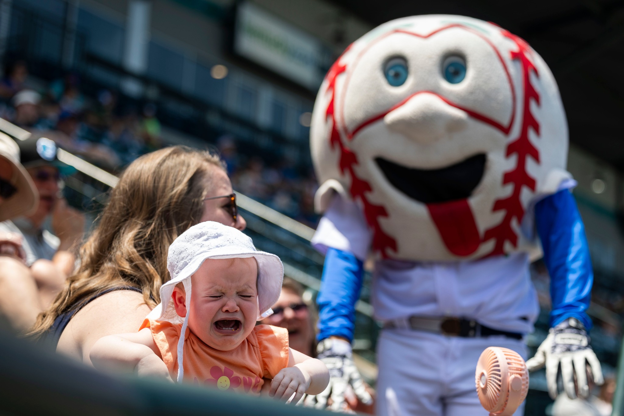 Crying baby sits near a smiling baseball mascot and stadium spectators.