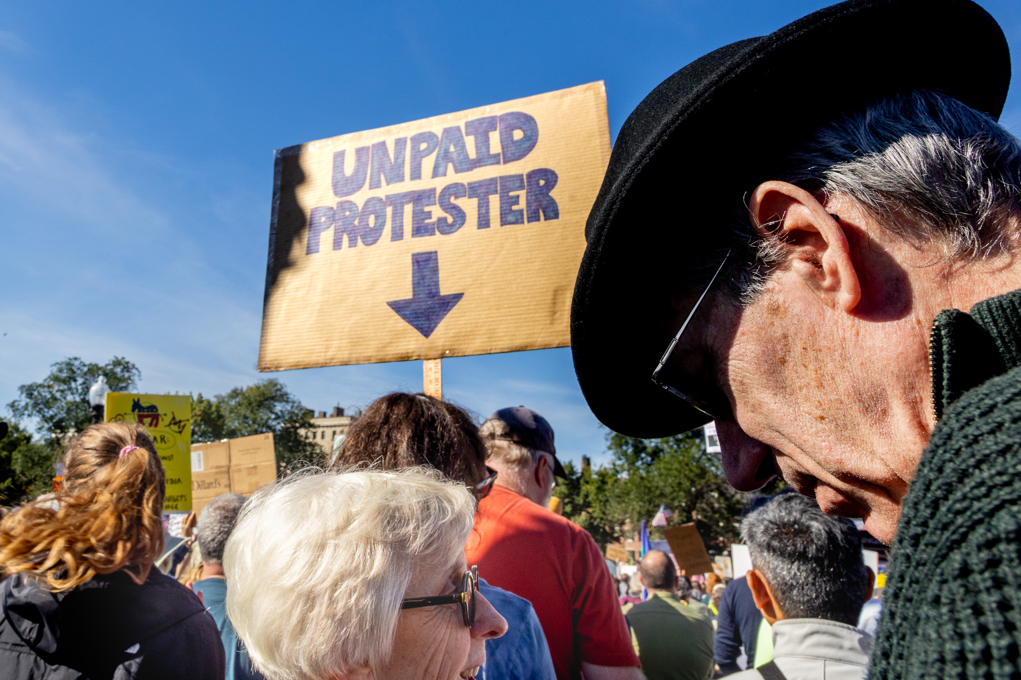 Crowd gathers beneath an Unpaid Protester sign as two people converse in the foreground.