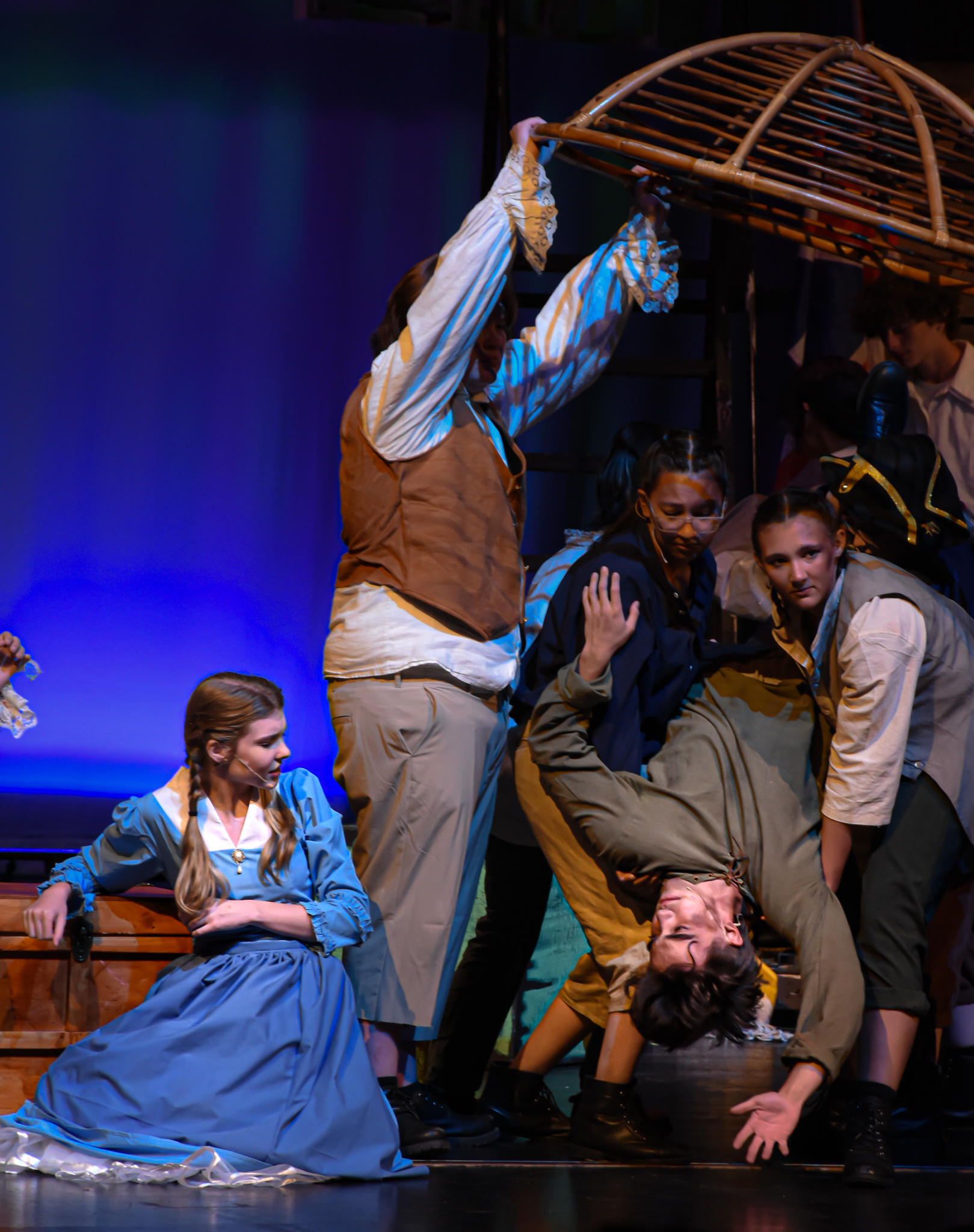 Costumed stage performers cluster beneath a wooden cage as one hangs upside down.