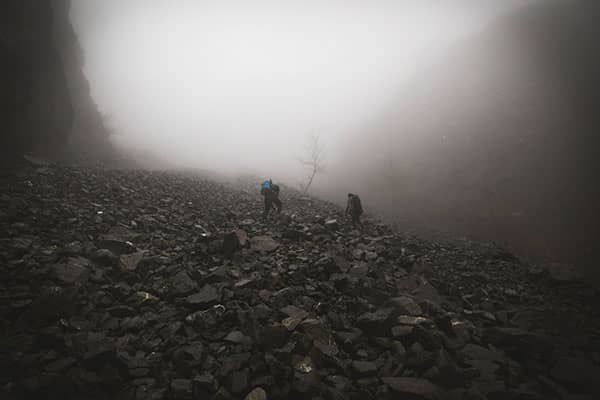 Two hikers cross a foggy, rock-strewn mountainside between steep cliffs.