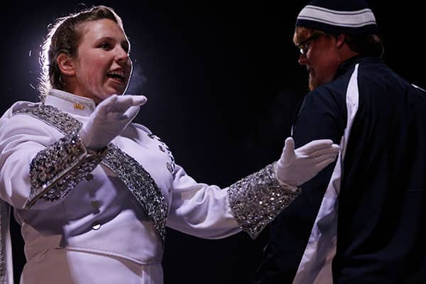 Uniformed marching band performers gesture to each other under stage lighting.