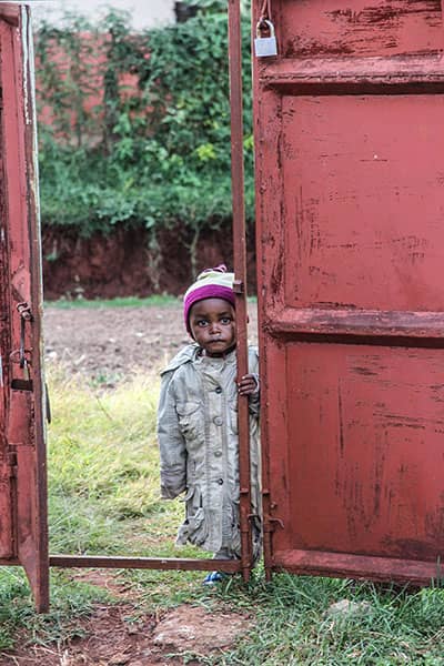 Young child in a coat and knit cap stands between weathered red gates.