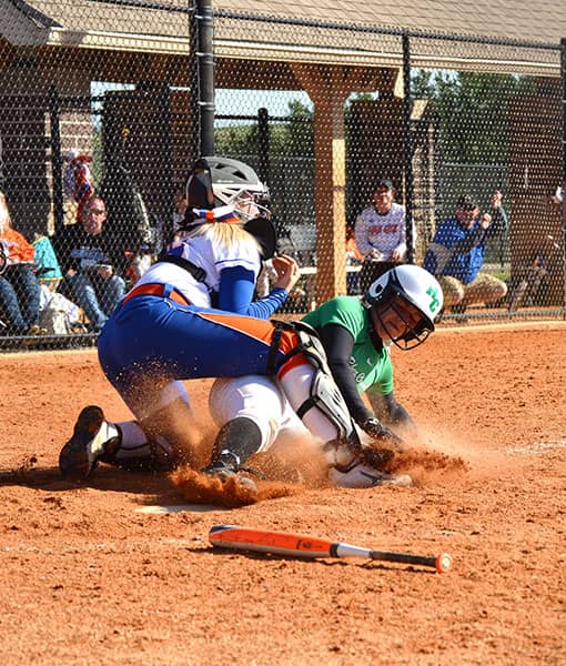 Softball catcher tags a sliding runner at home plate as dirt sprays.