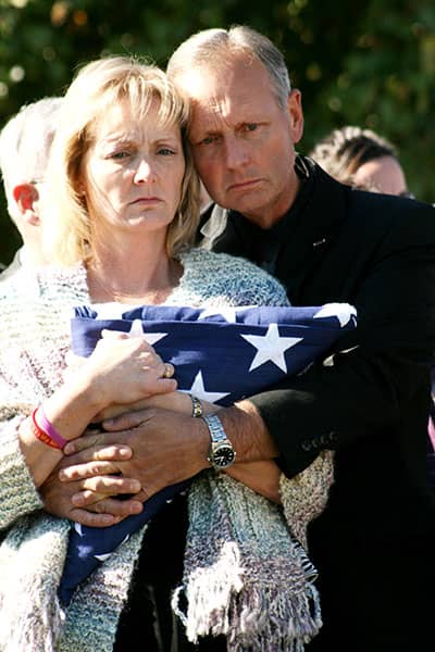 Two adults embrace while holding a folded American flag at an outdoor gathering.