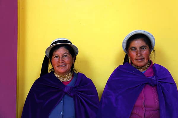 Two women wearing purple shawls and brimmed hats stand against a yellow wall.