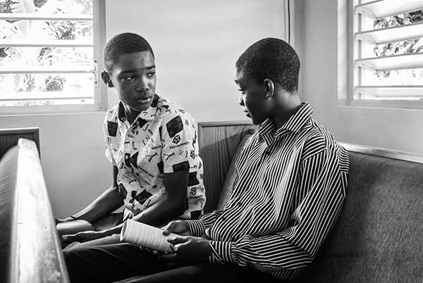 Two boys sit together in a church pew, one holding the other's bandaged hand.