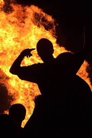 A silhouetted person gestures in front of a towering orange blaze at night.