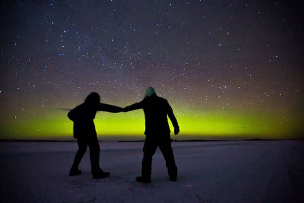 Two silhouetted figures hold hands beneath stars and green northern lights.