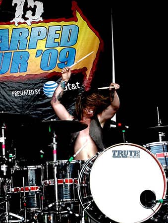 A shirtless drummer raises both sticks behind a drum kit during a live performance.