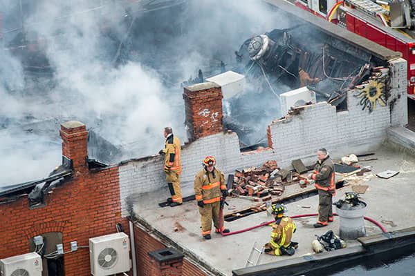 Firefighters stand on a damaged rooftop amid bricks, hoses, and heavy smoke.