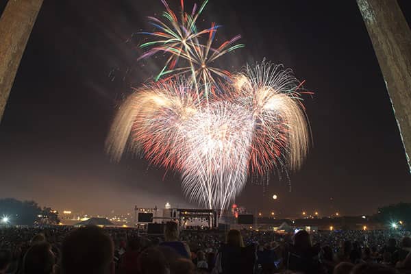 Colorful fireworks burst above a large nighttime crowd gathered near a distant stage.