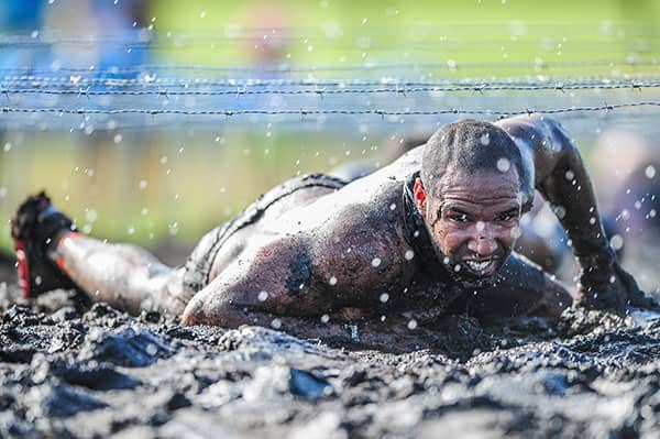 Mud-covered competitor crawls beneath barbed wire while water droplets fill the air.