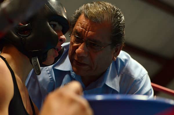 A coach leans close to a headgear-wearing boxer seated beside blue ring ropes.
