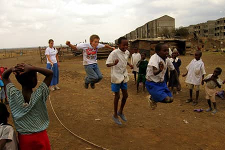 Children jump rope on a dirt field beside weathered concrete buildings.