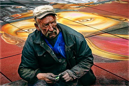 A seated man holds small objects before a brightly painted mural of faces.