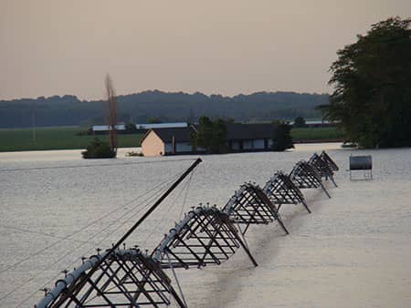 Floodwater surrounds farm buildings and a long row of partially submerged irrigation equipment.