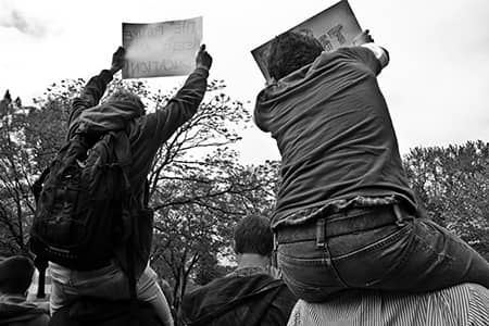 Two seated demonstrators raise signs above a crowd in a black-and-white park scene.
