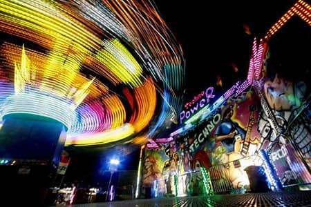 Spinning carnival ride lights streak beside a brightly illuminated funhouse at night.
