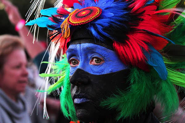 Person with vivid blue and black face paint wears a multicolored feathered headdress.