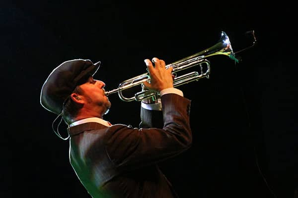 Trumpet player in a dark suit and cap performs under stage lighting.