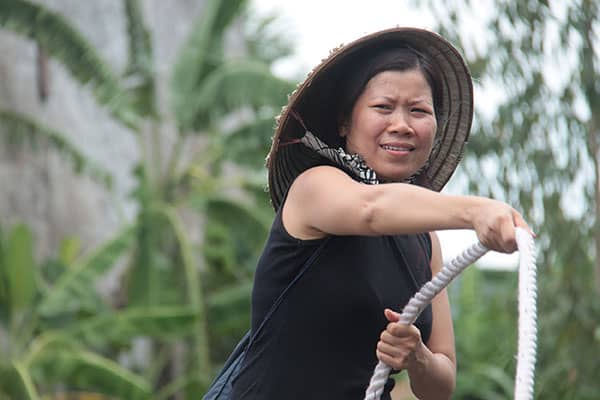 Woman in a conical hat pulls a thick rope amid tropical greenery.