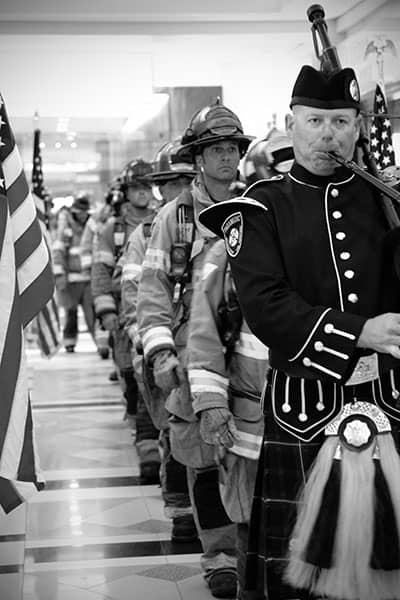 A bagpiper leads firefighters in procession through a flag-lined hallway.