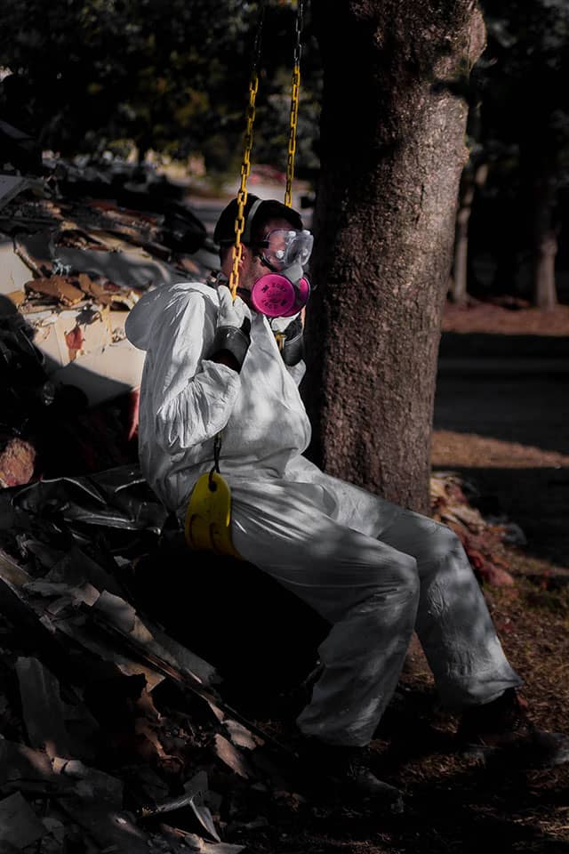 A person wearing protective coveralls and a respirator sits amid debris beside a tree.