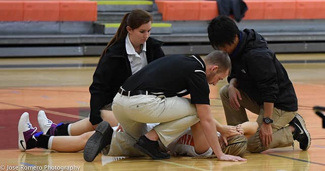 Three people kneel around an injured basketball player lying on a gym floor.