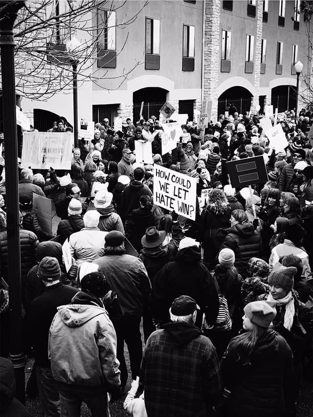 A dense street crowd carries protest signs, including one asking how hate could win.