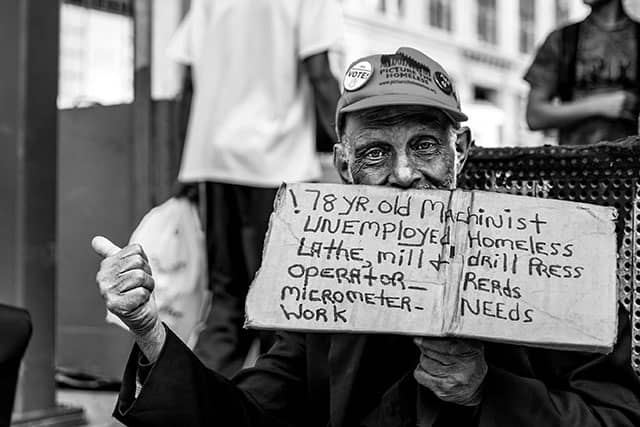 An older man holds a handwritten sign listing his machinist experience and employment needs.