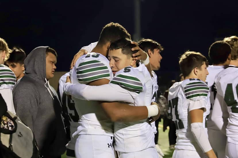 Two football players embrace on the field as teammates gather around them after a game.