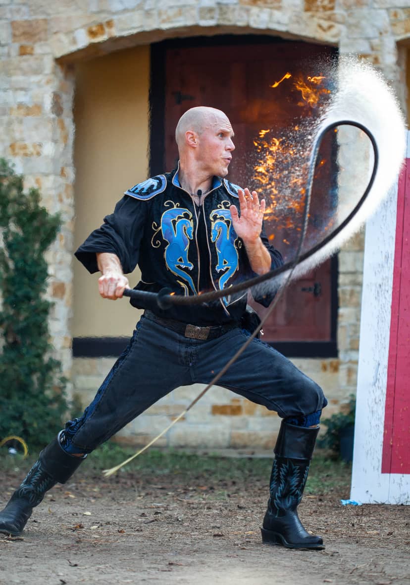 A whip performer swings a flaming loop while standing outside a stone building.