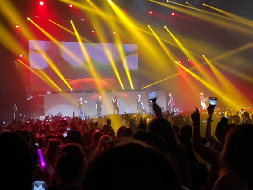 Performers sing beneath red and yellow stage lights before a crowded concert audience.