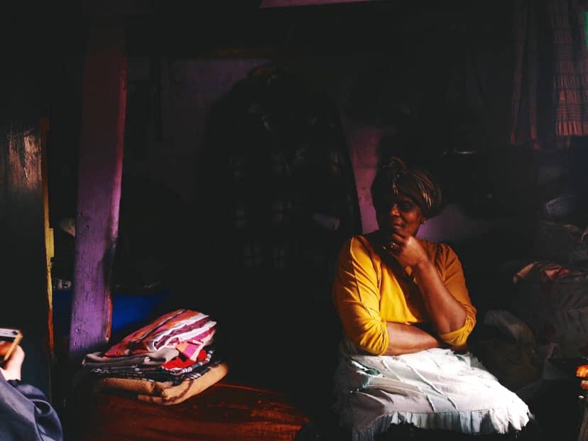 A seated woman in a yellow shirt rests beside folded fabrics in a dim room.