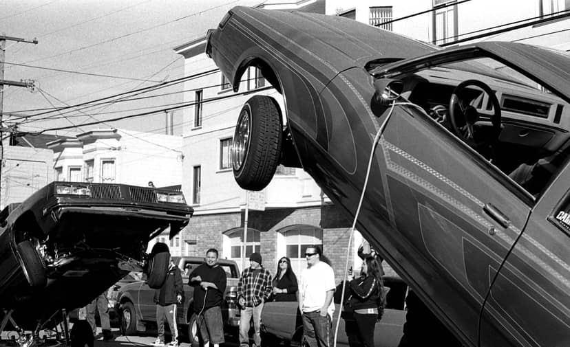 Two lowrider cars lift their front wheels above spectators on a city street.