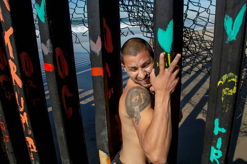 Shirtless tattooed man squeezes between painted metal border bars topped with tangled wire.