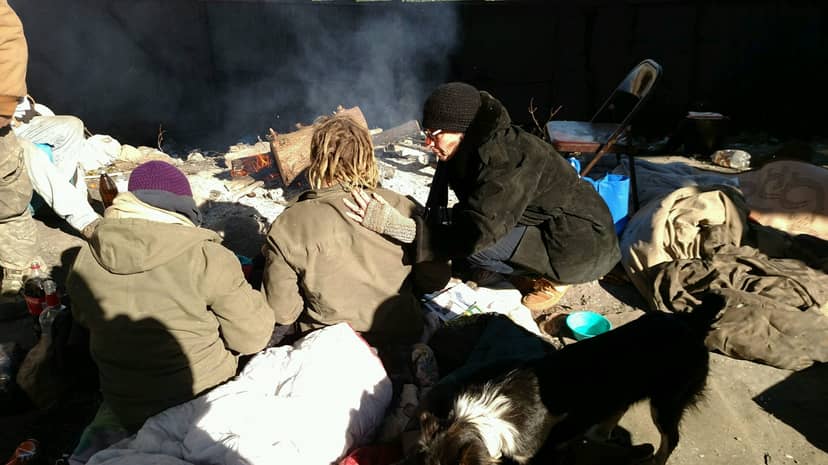 Three bundled people gather near a small outdoor fire with two dogs and scattered belongings.
