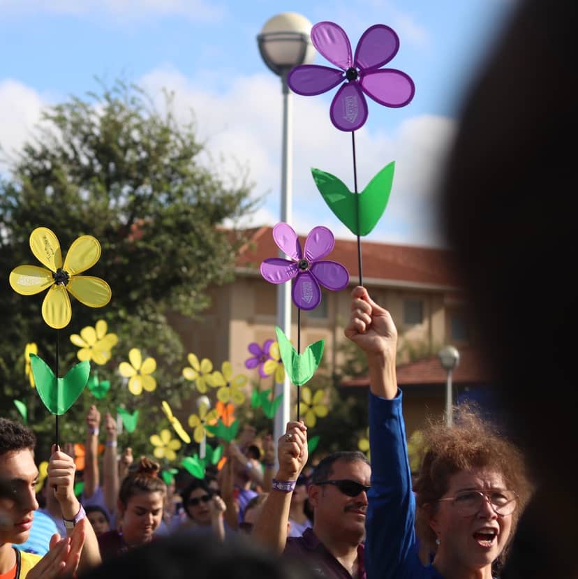 Crowd members hold colorful flower-shaped pinwheels aloft during an outdoor gathering.