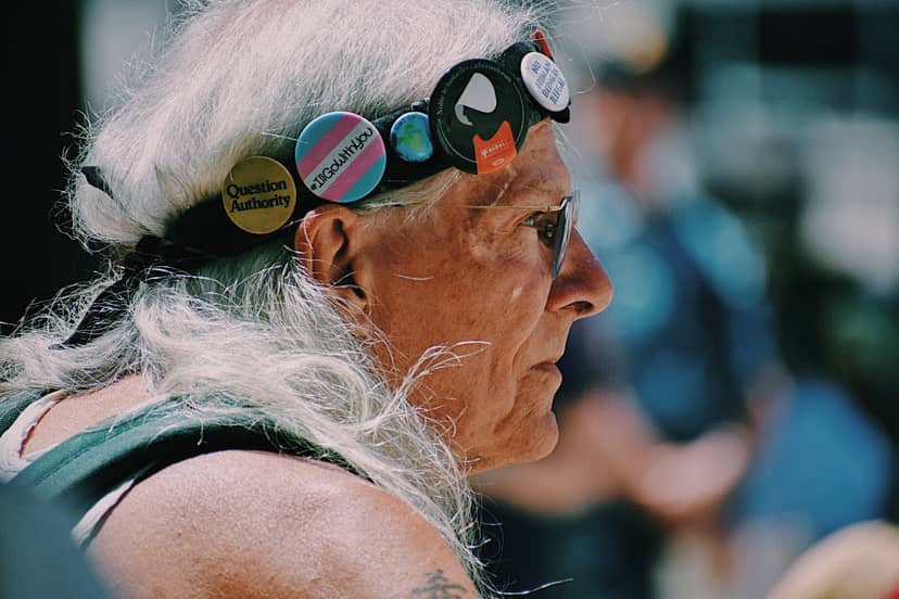 White-haired person in sunglasses wears a headband covered with political buttons and pins.