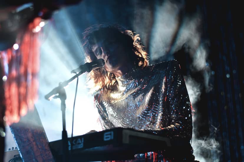 Backlit performer sings into a microphone while playing a keyboard on a smoky stage.